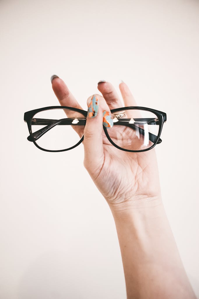 A hand with colorful nail art holding stylish eyeglasses against a light background.
